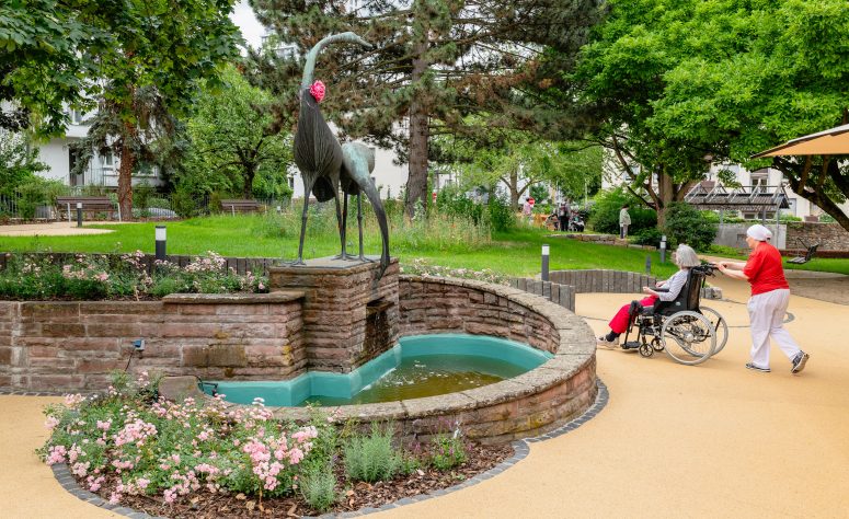 Eine Pflegerin fährt eine ältere Dame im Rollstuhl im Garten spazieren